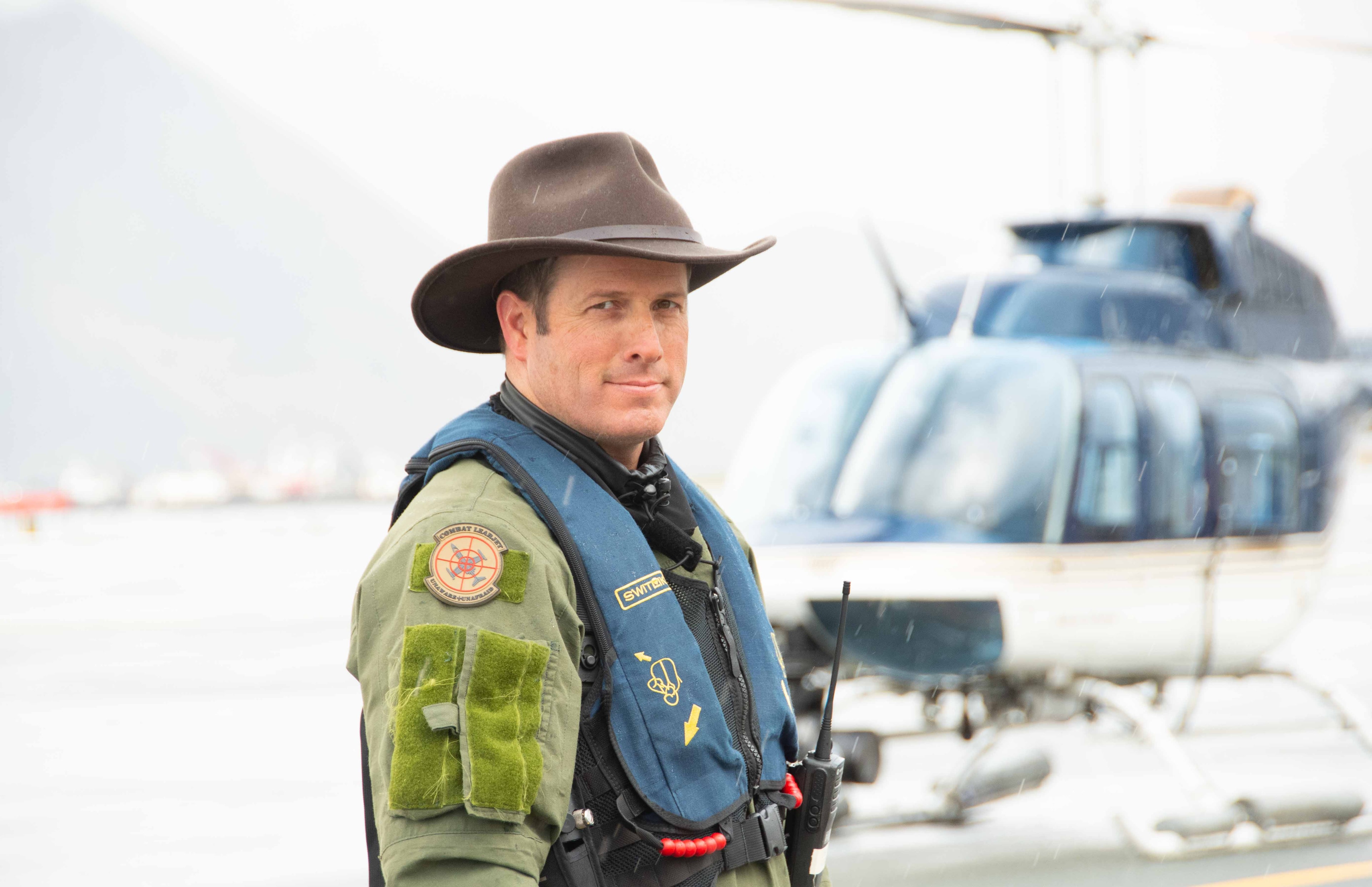 David Alan Arnold in survival vest and cowboy hat standing on dock in the rain, helicopter behind him, Deadliest Catch
